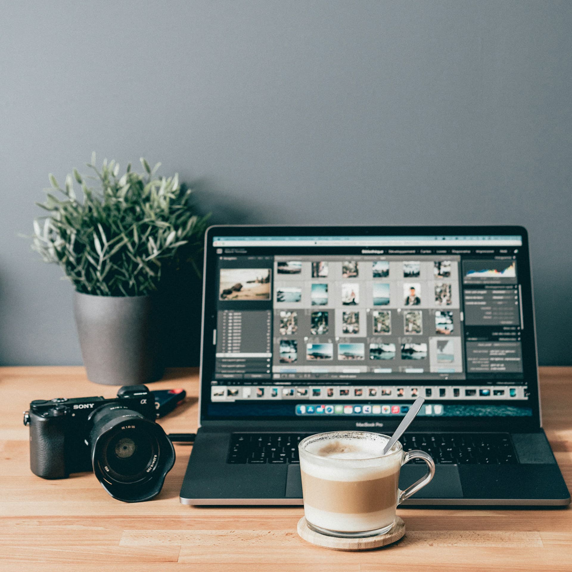 A MacBook running a photo editor with a mirrorless camera and a coffee on a wooden desk